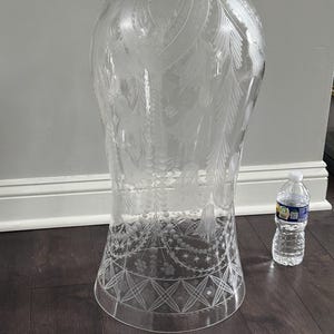 May include: A large, clear glass vase with intricate etched floral and feather designs. The vase has a wide base that tapers to a narrower midsection and flares out at the top. A water bottle is next to the vase for scale.
