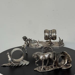 May include: A collection of silver napkin rings in various designs. The set includes rings shaped like a kangaroo on a fern, a horse-drawn carriage, a cherub, and a horse. The rings are displayed on a black surface.