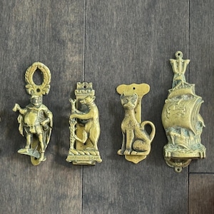 May include: A collection of antique brass door knockers in various designs, including a figure, a bear, a cat, and a ship. The door knockers are arranged on a dark wooden surface. A measuring tape is next to the door knockers.