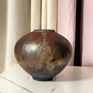 May include: A round, dark brown ceramic vase with a textured, metallic glaze. The glaze has shades of red, gold, and blue. The vase is sitting on a white marble surface.