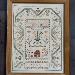 May include: A framed cross-stitch sampler with a white background and green, brown, and pink embroidery. The sampler features a house, flowers, and a border of stylized leaves. The text "M. Schultz 1940" is embroidered in the bottom right corner.