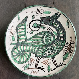 May include: A hand-painted ceramic plate featuring a stylized bird design in green and black against a white background. The bird has a long neck and tail, with leaf-like accents. The plate's rim is green.