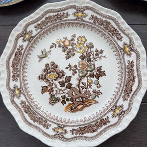 May include: A round ceramic plate with a scalloped edge featuring a central botanical illustration of flowers and a small butterfly in shades of yellow, green, and brown. The border has a brown floral pattern with yellow geometric accents.