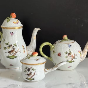 May include: A set of three white porcelain teaware pieces. The set includes a tall coffee pot, a round teapot, and a small creamer. Each piece is decorated with bird and butterfly motifs, gold trim, and a peach-colored knob.