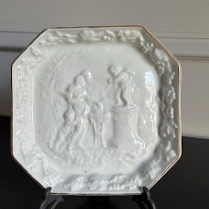 May include: White octagonal decorative plate with a raised relief design featuring classical figures. The plate has a gold trim and is displayed on a black stand. The relief depicts a scene with multiple figures and architectural elements.