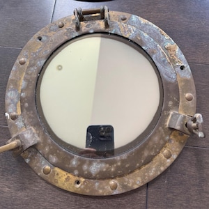 May include: A round, antique brass porthole with a glass window. The porthole has a weathered appearance with visible rust and corrosion. It features a hinged top and a locking mechanism on the side, suggesting a nautical theme.