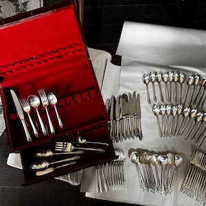 May include: A large silver flatware set featuring ornate floral handles, including knives, forks, and spoons. Some pieces are arranged inside a dark wooden storage chest with a vibrant red velvet interior, while others are laid out on white paper.