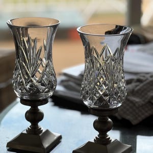 May include: Two clear glass candle holders with intricate cut crystal designs. The holders sit atop bronze-colored pedestals with a square base and a round, bulbous center. The glass has a flared top, and the background is blurred.