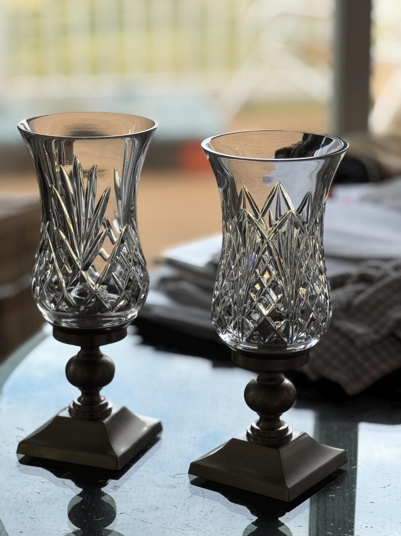 May include: Two clear glass candle holders with intricate cut crystal designs. The holders sit atop bronze-colored pedestals with a square base and a round, bulbous center. The glass has a flared top, and the background is blurred.