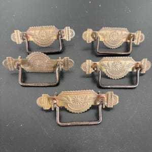 May include: Five antique brass drawer pulls with ornate floral and sunburst designs. Each pull has a rectangular backplate with a curved handle. The pulls are aged with a dark patina, suggesting vintage use.