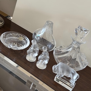 May include: A collection of clear glass decorative items, including a fish-shaped dish, two small figurines, a pear-shaped vase, a decorative bottle, and a bison figurine. The items are arranged on a dark brown surface.
