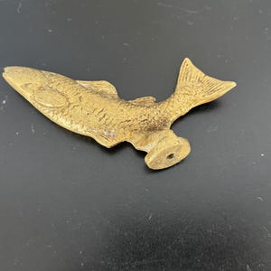 May include: A gold-colored metal fish figurine with a detailed design. The fish is in a jumping position with its tail raised. The figurine has a small hole in the bottom, suggesting it may be a bottle stopper.