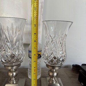 May include: Two clear crystal candle holders with a diamond pattern, each approximately 12 inches tall, sit on a brushed silver base. The candle holders have a rounded base and a square base. A measuring tape is placed between the candle holders.