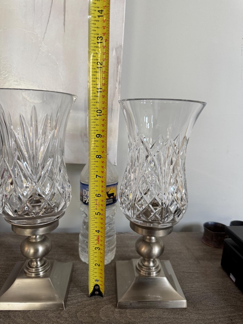 May include: Two clear crystal candle holders with a diamond pattern, each approximately 12 inches tall, sit on a brushed silver base. The candle holders have a rounded base and a square base. A measuring tape is placed between the candle holders.