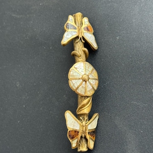 May include: A gold-toned decorative pin featuring a stylized design of butterflies and a flower. The butterflies have white and brown accents. The flower has a mosaic-like pattern. The pin is approximately 3 inches long.
