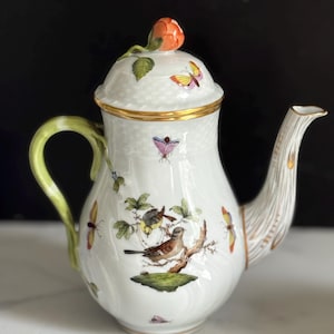 May include: White porcelain teapot with a lid topped with an orange rose. The teapot features a green handle, a textured spout, and is decorated with butterflies and birds. Gold trim accents the lid and base.