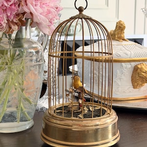May include: A vintage-style brass birdcage featuring a domed top with a circular hanging loop. Inside the cage, a small yellow and brown bird figurine is perched on a branch with tiny leaves, set upon a dark circular base with a brass rim.