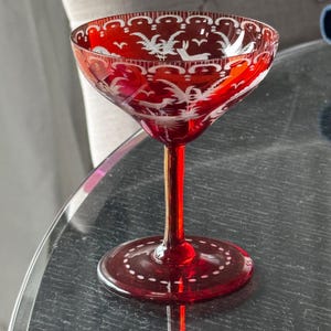 May include: A ruby red glass coupe with a slender stem and circular base. The bowl features etched white designs of deer and foliage, with a decorative scalloped border around the rim and a dotted pattern encircling the base of the glass.