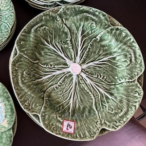 May include: A round ceramic plate shaped like a green cabbage leaf with prominent white veins radiating from the center. The surface has a glossy finish and textured details mimicking natural foliage. A small rectangular label with a rooster icon is attached.