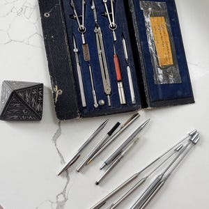 May include: A vintage black leather case with a blue interior containing a set of drafting tools, including compasses, rulers, and a pencil. The case is open and the tools are visible. There are also several silver pens and a small black pyramid on a white surface.