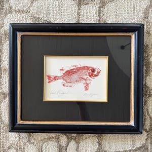 May include: Framed art print featuring a red fish illustration. The artwork is set within a black frame with a gold inner border. The fish is depicted in detail, with visible scales and fins. The print is signed by the artist.