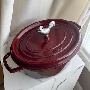May include: A burgundy oval Staub Dutch oven with a silver rooster-shaped handle. The pot has two side handles and the brand name is embossed on the side. The pot is sitting on a white surface.