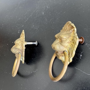 May include: Two antique brass lion head drawer pulls. Each pull features a detailed lion's head with a ring in its mouth. The pulls are mounted on a dark surface, showcasing their aged, weathered appearance.