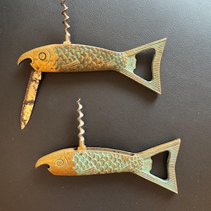 May include: Two bronze-colored fish-shaped bottle openers with a corkscrew and a small knife. The fish have a textured scale pattern and a green patina. The openers are on a dark surface.