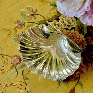 May include: A silver-colored, shell-shaped dish with a ridged design. The dish is on a yellow surface with floral painted accents and is next to a bouquet of flowers.