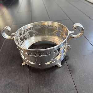 May include: A silver-colored metal bowl with ornate cut-out designs and two curved handles. The bowl has three small feet and a circular shape, suitable for serving or display.