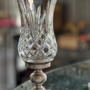 May include: A clear crystal glass candle holder with a diamond pattern and a silver-colored metal base. The candle holder has a flared top and a round, ball-shaped center. The base is square.
