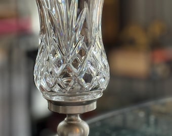 May include: A clear crystal glass candle holder with a diamond pattern and a silver-colored metal base. The candle holder has a flared top and a round, ball-shaped center. The base is square.