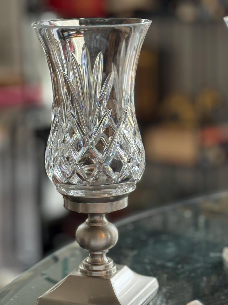 May include: A clear crystal glass candle holder with a diamond pattern and a silver-colored metal base. The candle holder has a flared top and a round, ball-shaped center. The base is square.