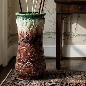 May include: A tall, cylindrical ceramic umbrella stand featuring a glossy brown glaze on the lower body and a vibrant green glaze on the flared rim. The exterior is decorated with intricate, raised relief scrollwork and floral motifs.