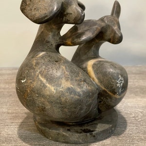 May include: A black and gray stone sculpture of two stylized animals, possibly dogs, with their heads touching. The sculpture is on a round base.