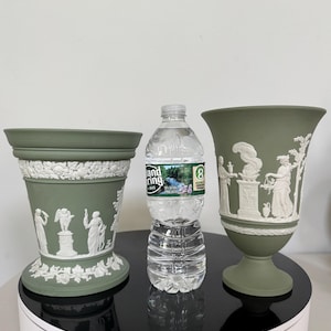 May include: Two sage green ceramic vases with white relief designs of classical figures and foliage. The vase on the left is a planter style, and the vase on the right is a pedestal vase. A water bottle is between the vases.