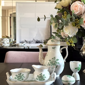 May include: A white porcelain tea set with green floral designs, including a teapot, creamer, sugar bowl, teacup, and serving tray. The set is arranged on a dark table with a bouquet of flowers in the background.