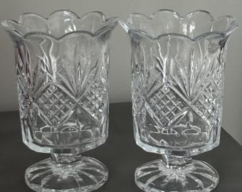May include: Two clear glass candle holders with a scalloped rim and diamond-cut pattern. The candle holders have a pedestal base and are designed to hold candles. The glass is clear and reflects light, creating a sparkling effect.