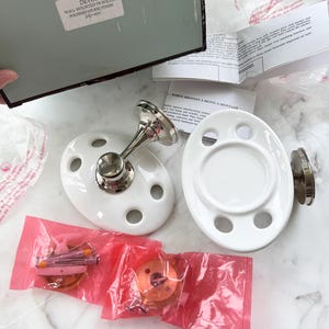 May include: Two white ceramic toothbrush holders with four slots each. The holders are attached to silver metal wall mounts. The holders are packaged in pink plastic bags with hardware.
