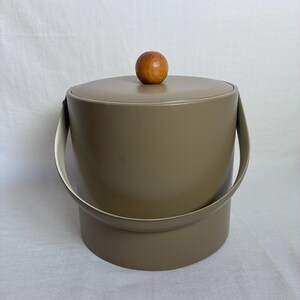 May include: A cylindrical taupe ice bucket with a smooth matte finish, featuring a matching lid topped with a round wooden knob handle and a curved metal swing handle attached to the sides of the container for easy carrying and storage.