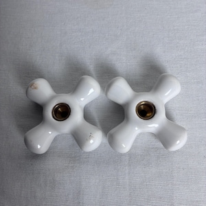 May include: Two white ceramic cross-shaped faucet handles with a smooth, glossy finish. Each handle features a central circular brass-colored metal insert with a threaded opening, designed for attachment to a plumbing fixture valve stem.