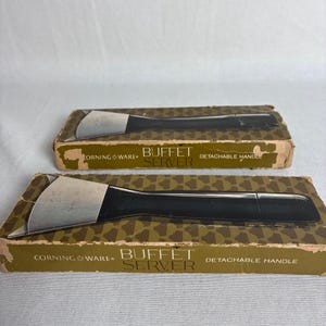 May include: Two Corning Ware Buffet Server detachable handles, each featuring a black plastic grip and a silver-colored metal attachment end. The handles are displayed on their original rectangular cardboard boxes with brown and green patterns.