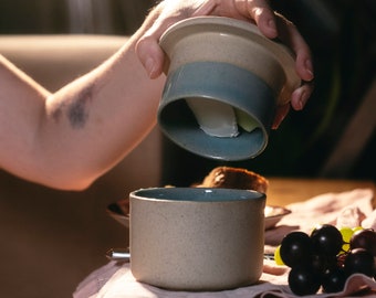 May include: A two-piece ceramic butter crock with a speckled beige exterior and a smooth blue-grey interior. The cylindrical base holds the butter, while the inverted lid features a deep well for water, designed to keep the butter fresh and spreadable.