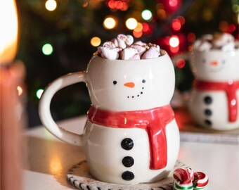 May include: A white ceramic mug shaped like a snowman with a red scarf, black dot eyes, a small orange carrot nose, and three black buttons on the front. The mug is filled with small marshmallows and sits on a round, white, braided coaster.