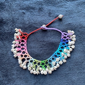 May include: A colorful beaded necklace with a rainbow gradient design. The necklace features a series of small, silver-colored charms hanging from the bottom edge. The necklace is set against a dark blue background.