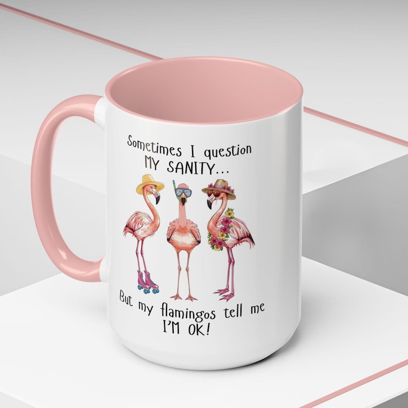 May include: White ceramic mug with a pink interior and handle. The mug features three flamingo illustrations wearing hats and accessories. Text on the mug reads "Sometimes I question my sanity... But my flamingos tell me I'M OK!"