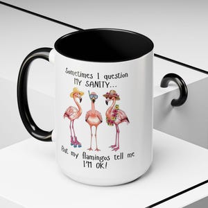 May include: White ceramic mug with a black interior and handle. The mug features a whimsical illustration of three pink flamingos wearing hats and accessories, with the text "Sometimes I question MY SANITY... But my flamingos tell me I'M OK!"