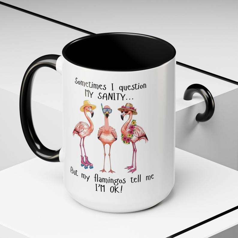 May include: White ceramic mug with a black interior and handle. The mug features a whimsical illustration of three pink flamingos wearing hats and accessories, with the text "Sometimes I question MY SANITY... But my flamingos tell me I'M OK!"