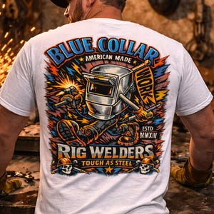 May include: White t-shirt with a colorful graphic featuring a welding scene. The design includes the words "BLUE COLLAR AMERICAN MADE WORK" and "RIG WELDERS TOUGH AS STEEL". The shirt has a crew neck and short sleeves.