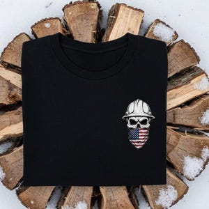 May include: Black t-shirt featuring a skull wearing a white construction helmet and an American flag bandana. The skull design is detailed with shading and highlights. The shirt is displayed on a background of wood and snow.